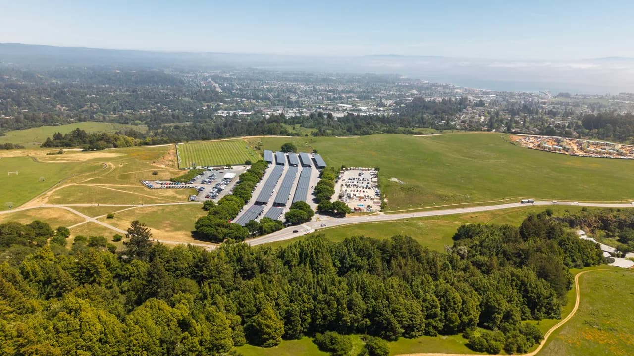 Rows of long structures sit on open green hills above a wooded slope and a distant city. Coast & City photo 9 of 11.